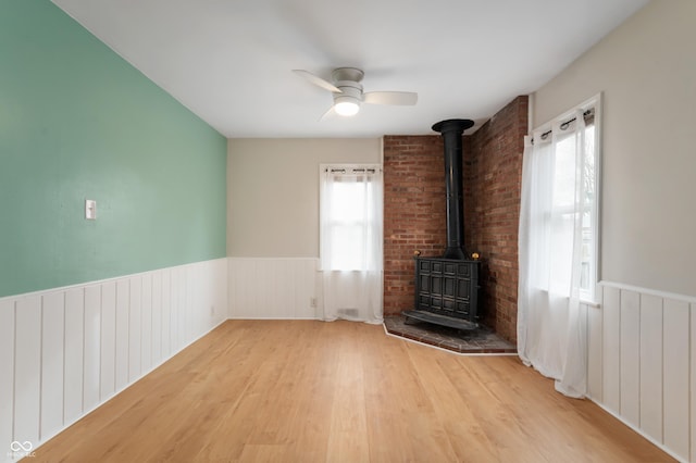 unfurnished living room with wainscoting, a wood stove, ceiling fan, and wood finished floors