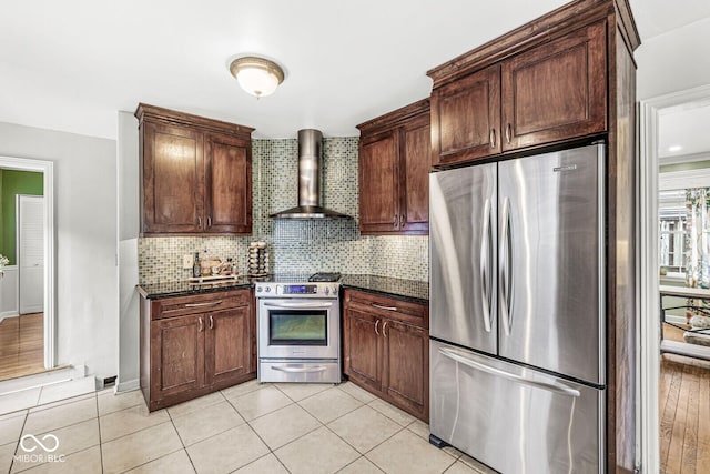 kitchen with light tile patterned floors, dark stone counters, stainless steel appliances, wall chimney exhaust hood, and tasteful backsplash