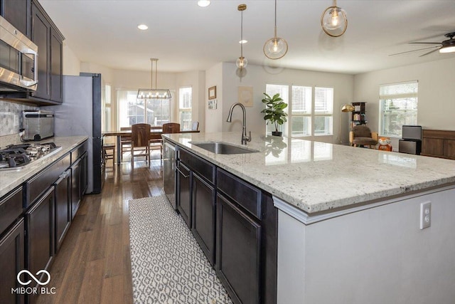 kitchen featuring a kitchen island with sink, a sink, stainless steel appliances, light stone countertops, and dark wood-style flooring