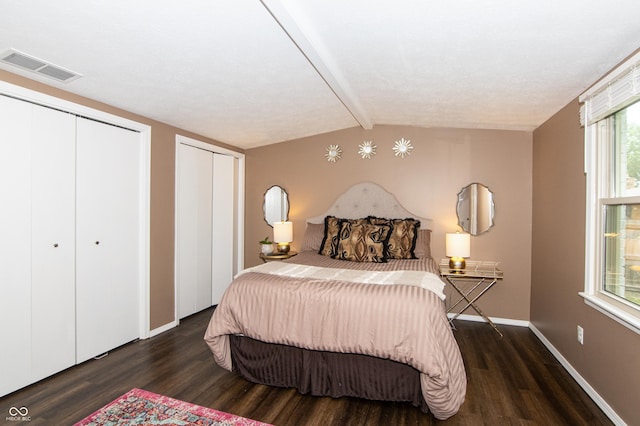 bedroom with wood finished floors, visible vents, baseboards, lofted ceiling with beams, and two closets