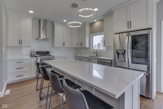 kitchen with light wood-type flooring, appliances with stainless steel finishes, a kitchen island, and wall chimney exhaust hood