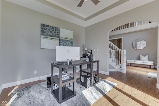 office space with a ceiling fan, wood finished floors, baseboards, a tray ceiling, and ornamental molding