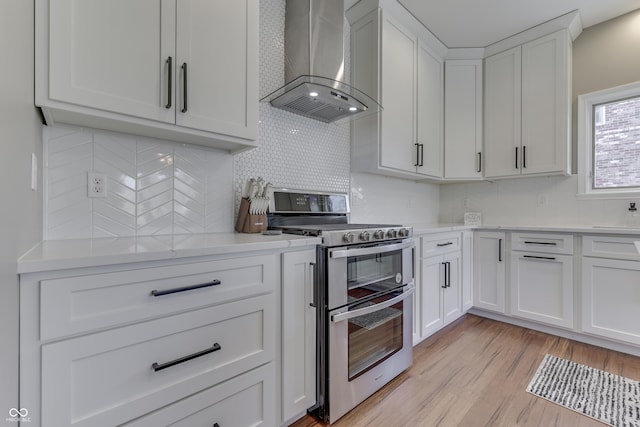 kitchen with double oven range, wall chimney range hood, white cabinets, light wood finished floors, and decorative backsplash