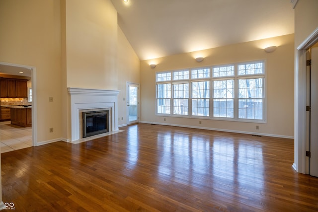 unfurnished living room featuring a fireplace with flush hearth, wood finished floors, baseboards, and high vaulted ceiling