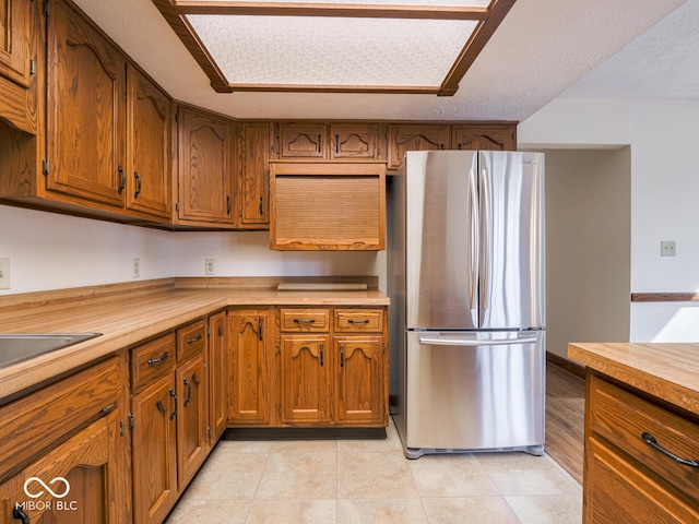 kitchen with a textured ceiling, freestanding refrigerator, brown cabinetry, light countertops, and light tile patterned floors