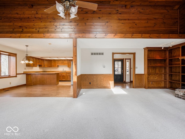 unfurnished living room with wooden walls, a ceiling fan, visible vents, wainscoting, and light carpet