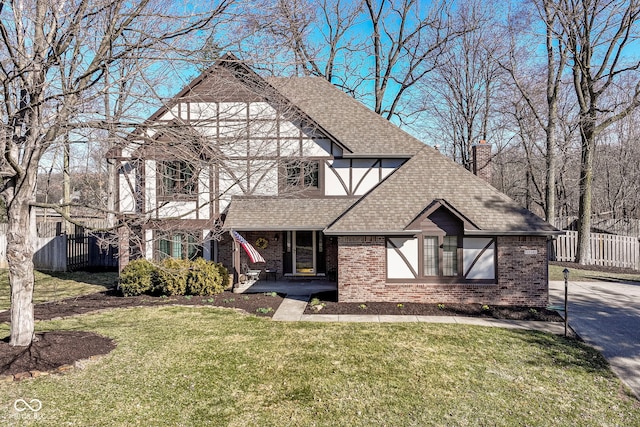 tudor-style house featuring fence, a front yard, a shingled roof, brick siding, and a chimney