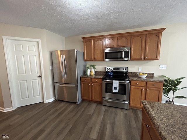 kitchen with brown cabinetry, stainless steel appliances, dark wood-type flooring, and dark stone counters