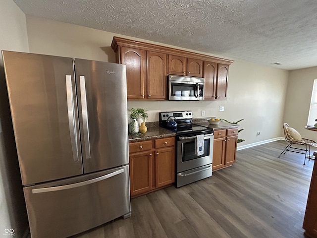 kitchen with a textured ceiling, dark wood finished floors, stainless steel appliances, brown cabinetry, and baseboards