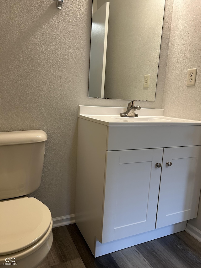 half bath with toilet, wood finished floors, baseboards, vanity, and a textured wall