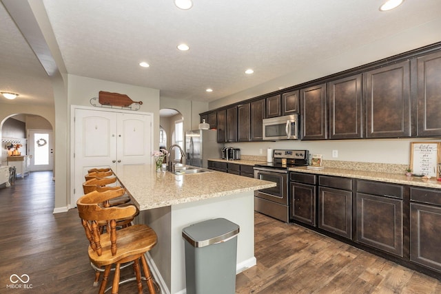 kitchen featuring dark brown cabinets, a breakfast bar, an island with sink, arched walkways, and stainless steel appliances