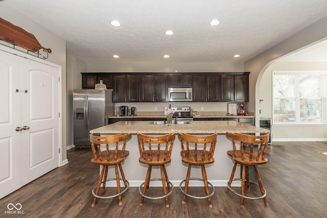 kitchen featuring a breakfast bar, a kitchen island with sink, stainless steel appliances, dark brown cabinetry, and dark wood-style flooring