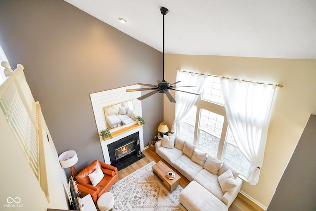 living room featuring wood finished floors, baseboards, high vaulted ceiling, a fireplace with flush hearth, and ceiling fan