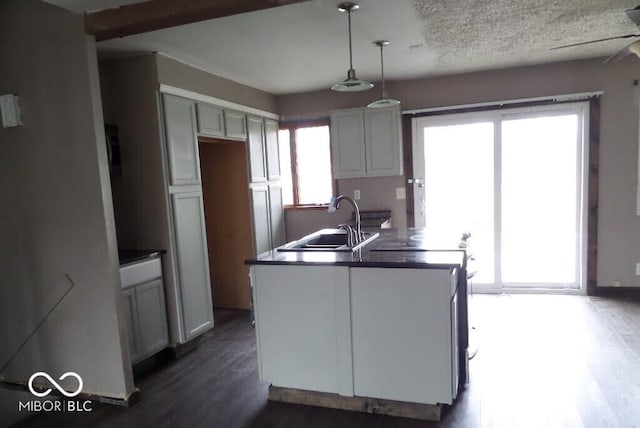 kitchen with decorative light fixtures, a sink, dark wood-type flooring, dark countertops, and an island with sink