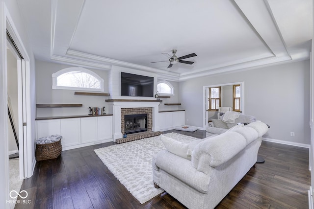 living area with a ceiling fan, a raised ceiling, a brick fireplace, baseboards, and dark wood-style flooring