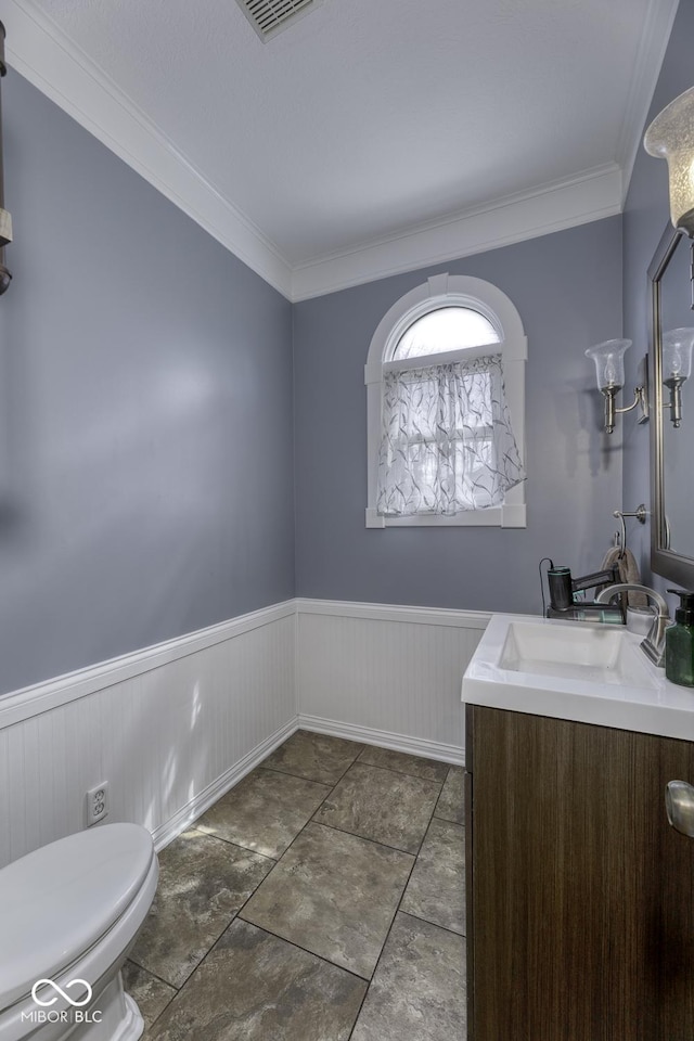 half bath featuring visible vents, crown molding, toilet, vanity, and wainscoting