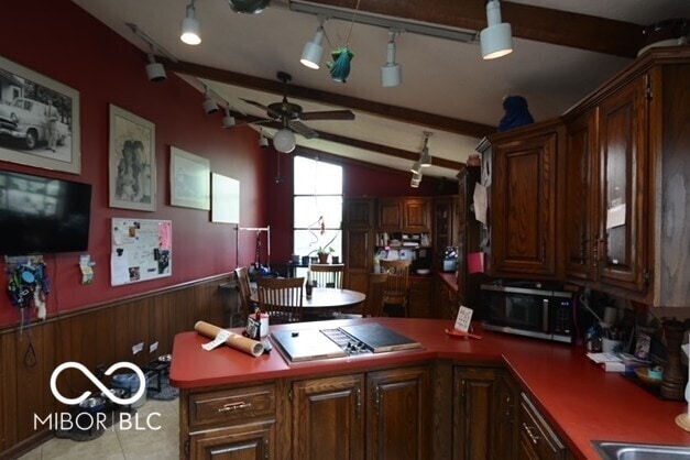 kitchen with vaulted ceiling with beams, stainless steel microwave, wainscoting, a ceiling fan, and a peninsula