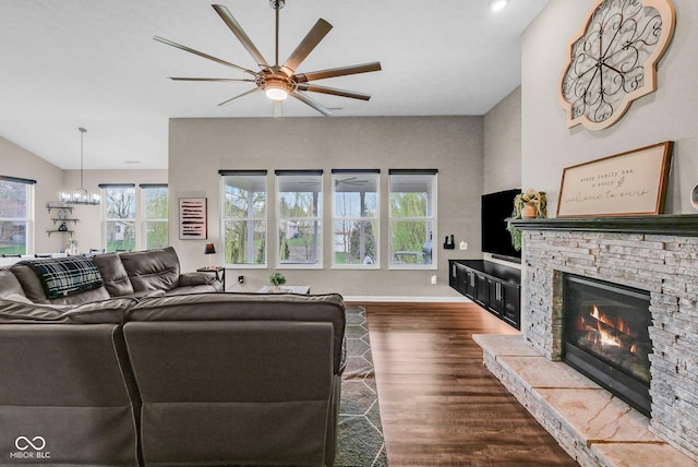living area featuring dark wood finished floors, a stone fireplace, baseboards, and ceiling fan with notable chandelier
