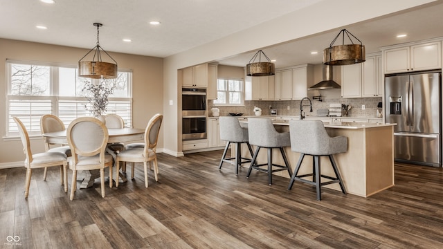 kitchen with light countertops, wall chimney exhaust hood, stainless steel appliances, an island with sink, and backsplash