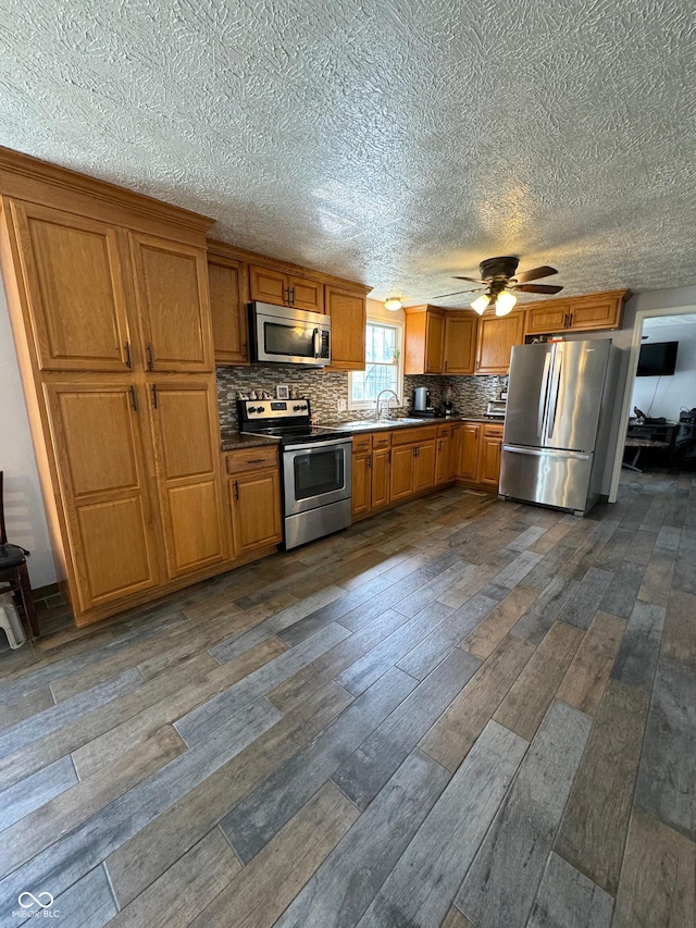 kitchen with appliances with stainless steel finishes, dark wood-type flooring, brown cabinets, ceiling fan, and a sink