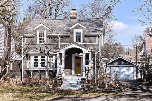 new england style home featuring a chimney, an outbuilding, a detached garage, aphalt driveway, and a shingled roof