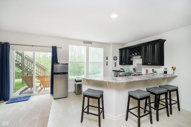 kitchen with a kitchen bar, light stone countertops, freestanding refrigerator, a peninsula, and dark cabinetry
