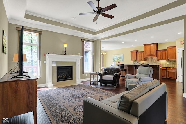 living room featuring dark wood-style flooring, a glass covered fireplace, ceiling fan with notable chandelier, a tray ceiling, and ornamental molding