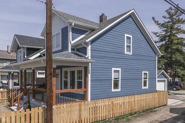 view of side of home featuring covered porch, a detached garage, roof with shingles, fence, and a chimney