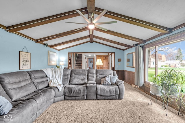 living room with a wainscoted wall, stairs, ceiling fan, carpet, and vaulted ceiling with beams