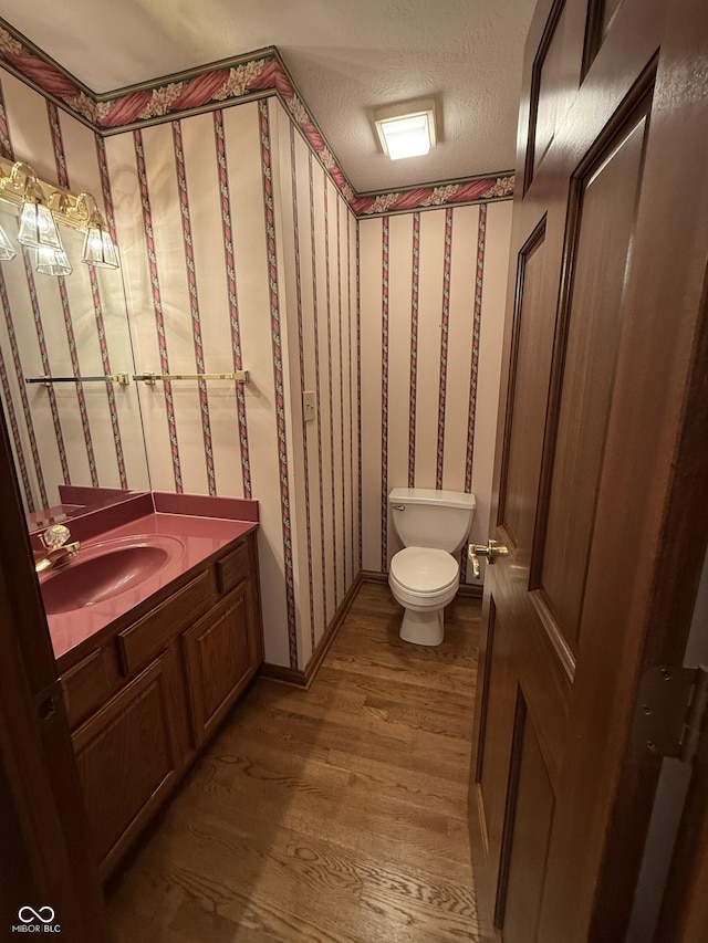 bathroom featuring a textured ceiling, wood finished floors, vanity, toilet, and wallpapered walls
