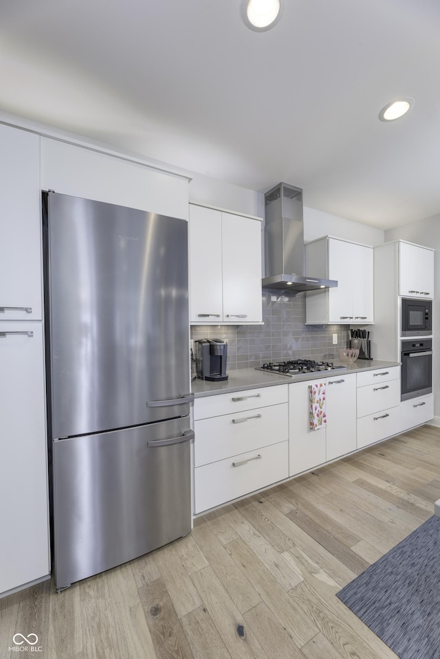 kitchen with wall chimney range hood, light wood-style flooring, stainless steel appliances, white cabinetry, and decorative backsplash