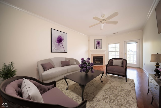 living area featuring ceiling fan, a tiled fireplace, wood finished floors, visible vents, and ornamental molding