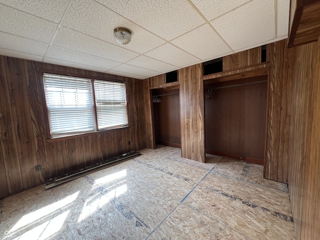 unfurnished bedroom with a baseboard radiator, wooden walls, a paneled ceiling, and multiple closets