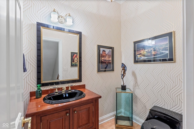 bathroom featuring baseboards, vanity, wallpapered walls, and wood finished floors