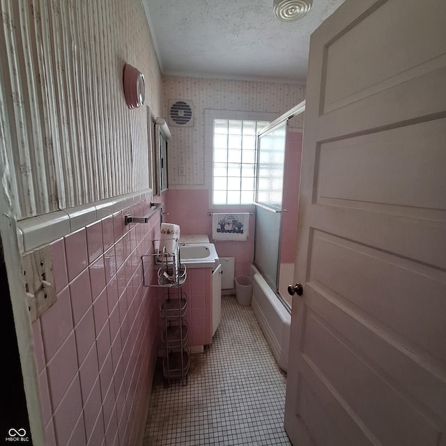 full bath with wallpapered walls, tile walls, tile patterned flooring, wainscoting, and bath / shower combo with glass door
