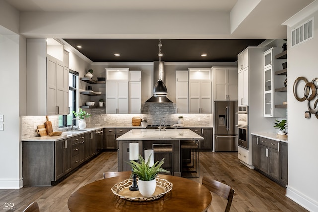 kitchen with open shelves, decorative backsplash, an island with sink, wall chimney range hood, and stainless steel appliances