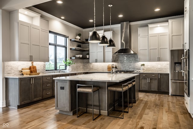 kitchen with wall chimney range hood, crown molding, open shelves, a center island with sink, and stainless steel appliances