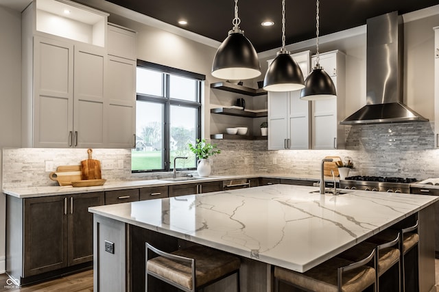 kitchen featuring wall chimney range hood, open shelves, a sink, a kitchen island with sink, and decorative backsplash