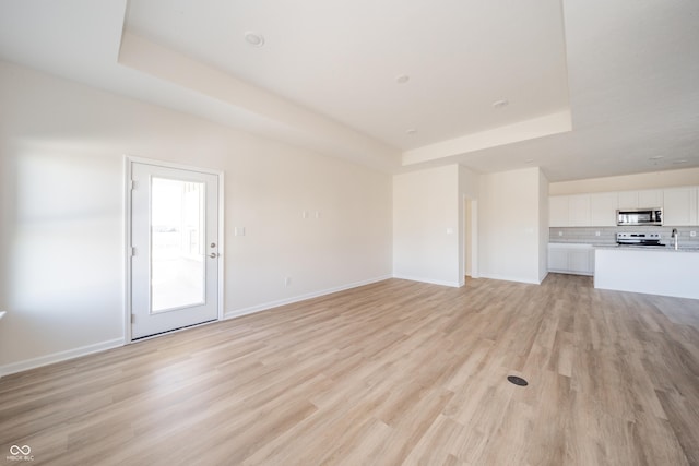 unfurnished living room with a sink, baseboards, a tray ceiling, and light wood finished floors