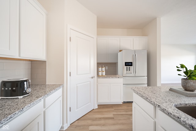 kitchen featuring light stone counters, white refrigerator with ice dispenser, light wood finished floors, white cabinetry, and backsplash
