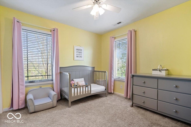 carpeted bedroom featuring a nursery area, a ceiling fan, and visible vents