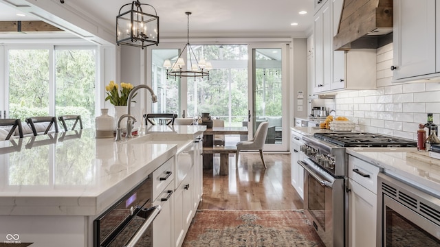 kitchen with wall chimney range hood, white cabinetry, stainless steel appliances, and a notable chandelier