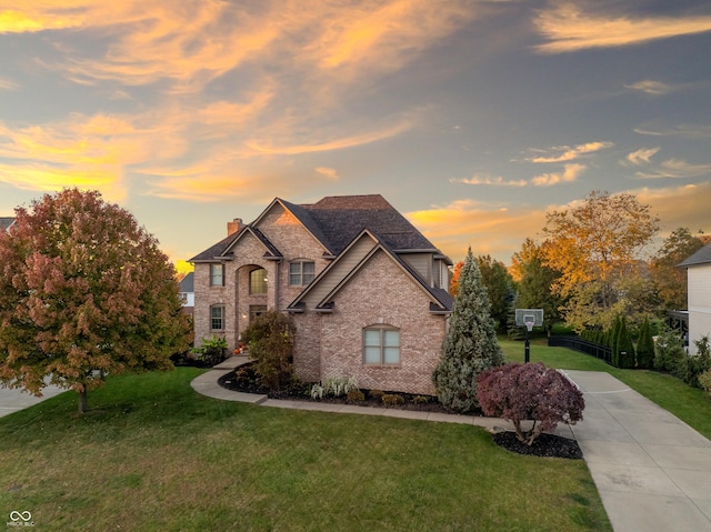 french country inspired facade featuring a lawn, driveway, a chimney, and brick siding