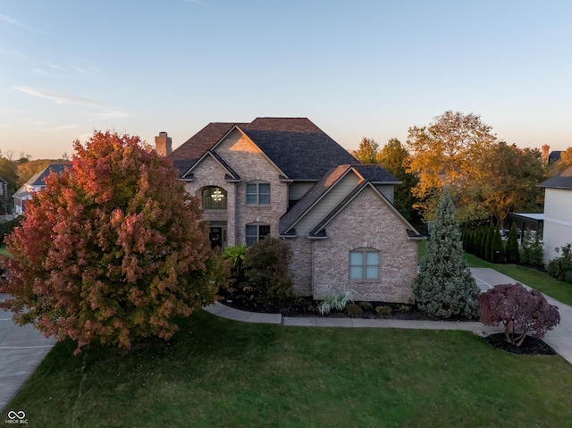 french country home with a shingled roof, a chimney, brick siding, and a yard