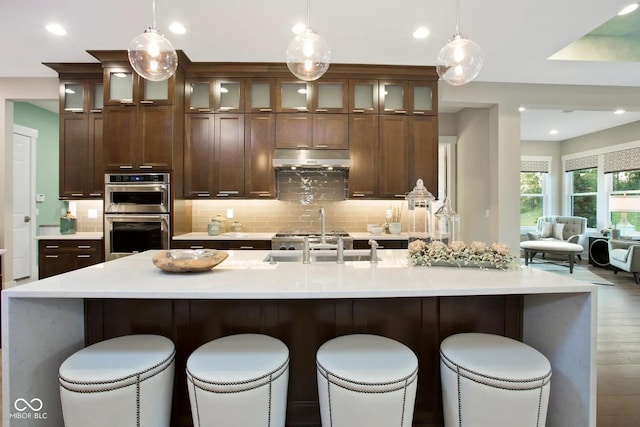 kitchen with a sink, stainless steel double oven, under cabinet range hood, light countertops, and tasteful backsplash
