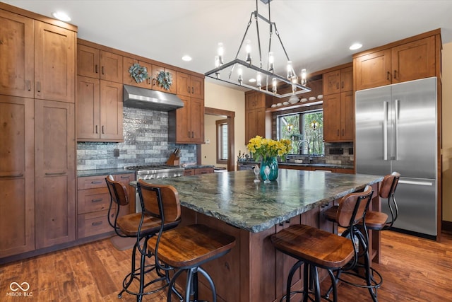 kitchen with built in refrigerator, under cabinet range hood, tasteful backsplash, recessed lighting, and brown cabinets
