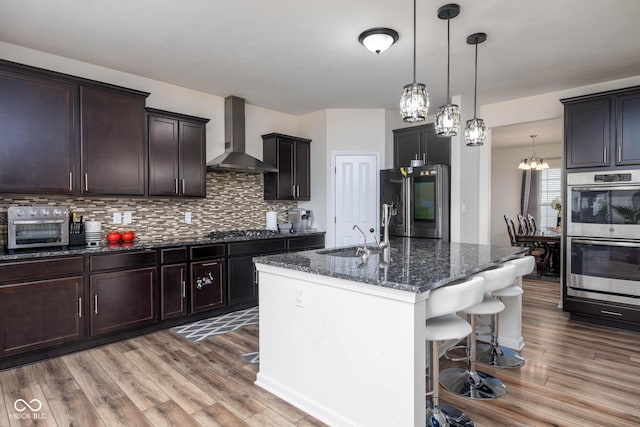 kitchen with dark stone countertops, a sink, a chandelier, wall chimney exhaust hood, and appliances with stainless steel finishes