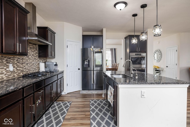 kitchen featuring stainless steel appliances, dark stone countertops, wood finished floors, a sink, and wall chimney exhaust hood