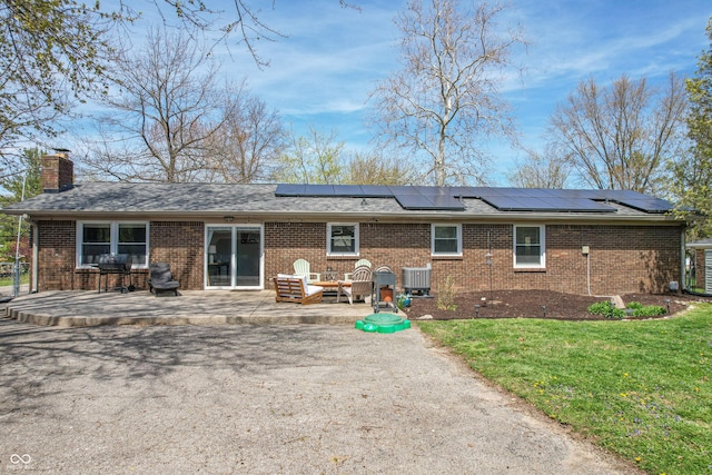 back of property with central AC unit, a chimney, brick siding, a lawn, and roof mounted solar panels