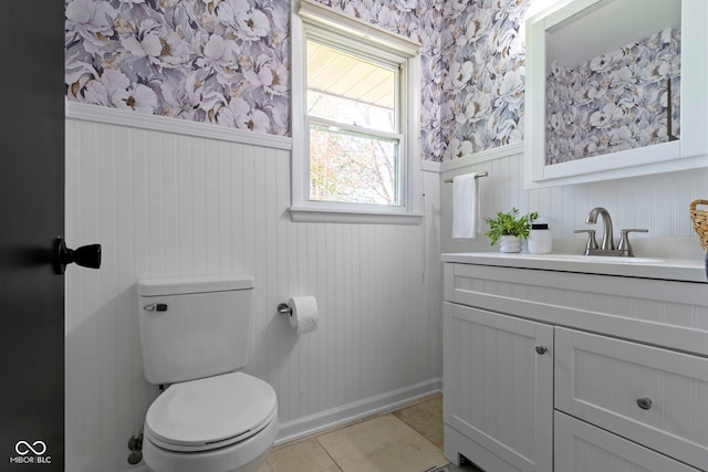 half bathroom featuring wallpapered walls, baseboards, toilet, vanity, and wainscoting